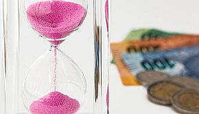 laufende Sanduhr mit Geldscheinen und Münzen im Hintergrund