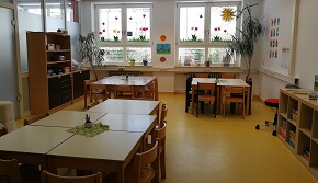 Ein Gruppenraum mit Tischen und Stühlen. Zusätzlich sind Spielregale auf der rechten und linken Seite.